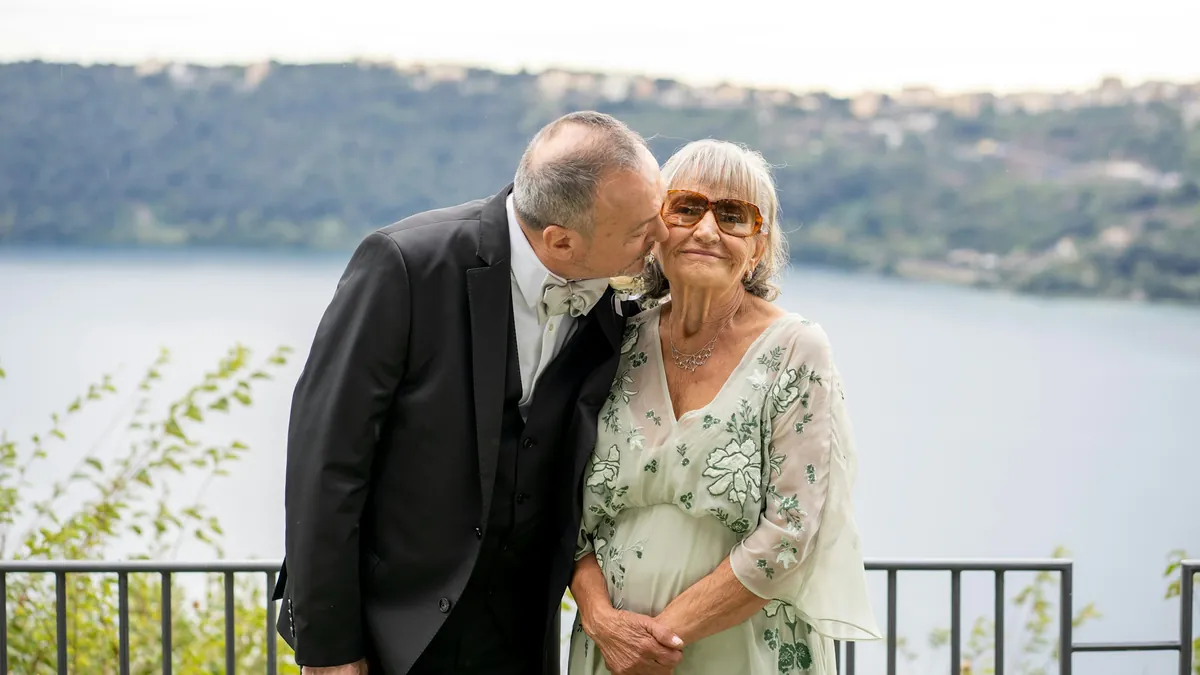 Él besa en la mejilla a su esposa, ambos vestidos de fiesta, frente a un lago