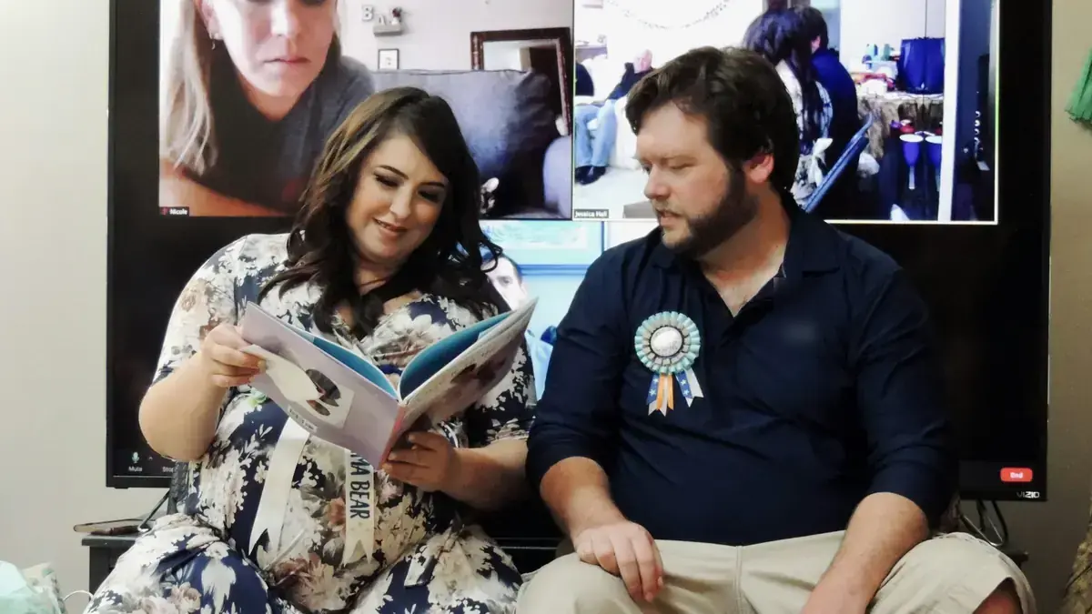 Una pareja lee un libro en su baby shower mientras la familia los acompaña por videollamada en la pantalla