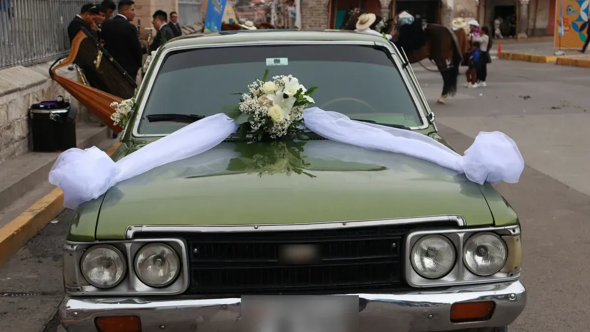 A wedding car decorated with flowers and white ribbons on a street in Ayacucho, Peru, with musicians and a horse behind