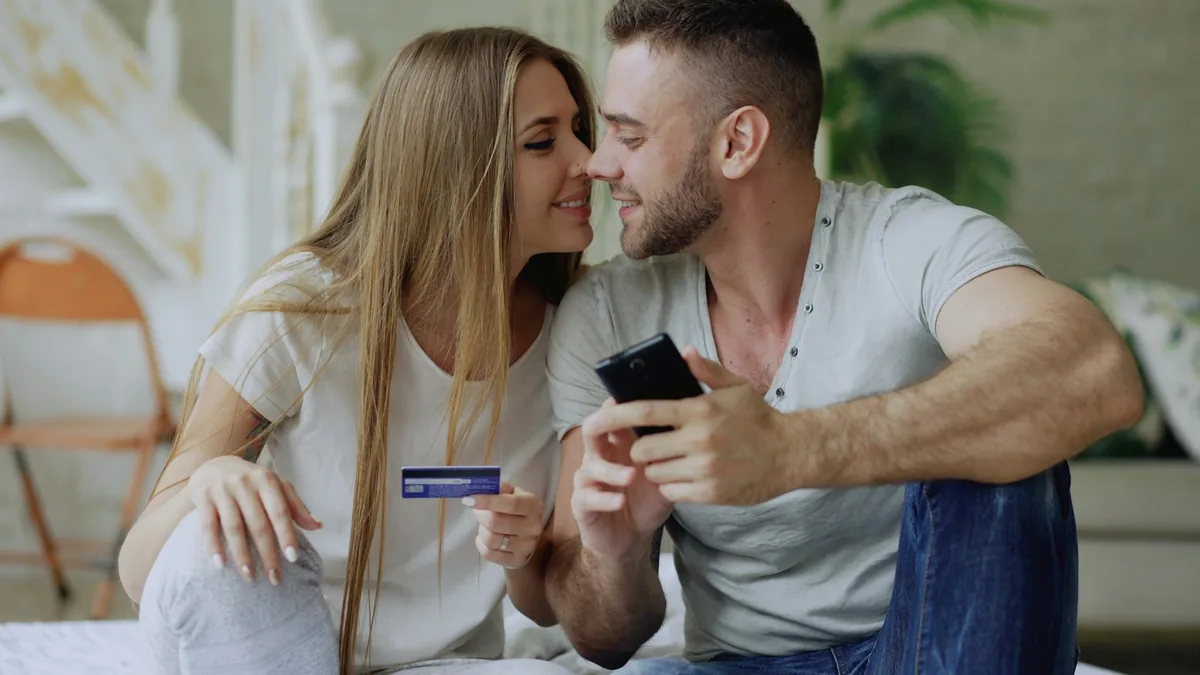 Una pareja sonríe en casa con una tarjeta bancaria y un celular en la mano