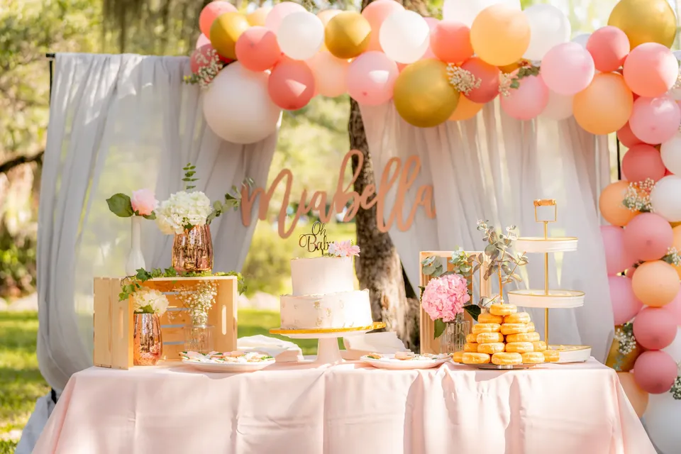 Mesa de baby shower con arco de globos, tarta y flores, montada en un jardín