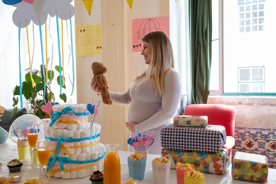 A mother-to-be smiles at a teddy bear at her baby shower, next to the diaper cake and the gifts