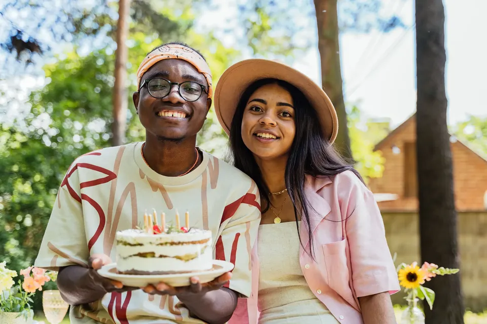 Una pareja sonríe sosteniendo una tarta de cumpleaños con velas encendidas, al aire libre
