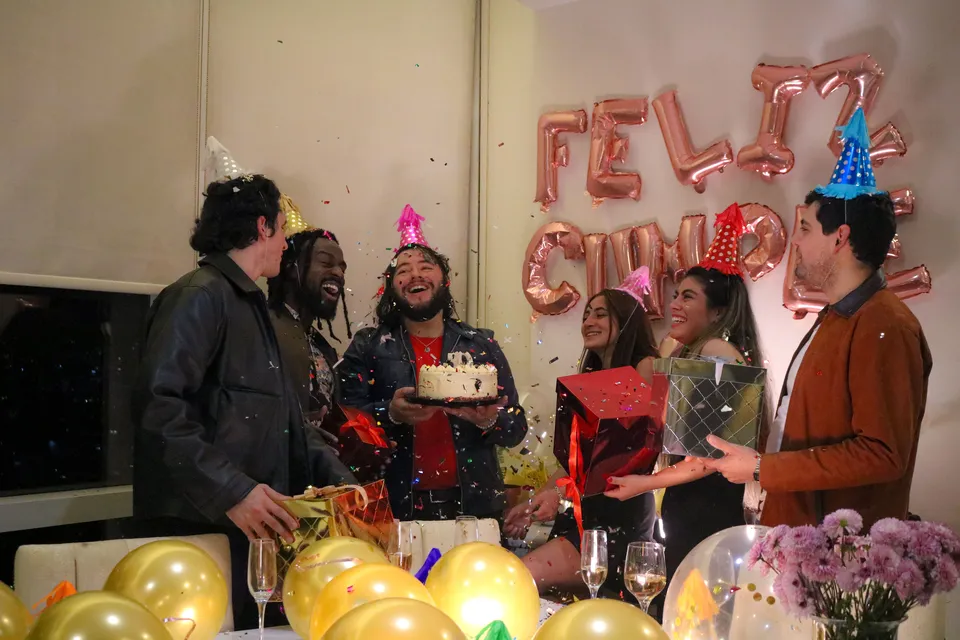 A group of friends celebrates a birthday with party hats, confetti and a cake with candles