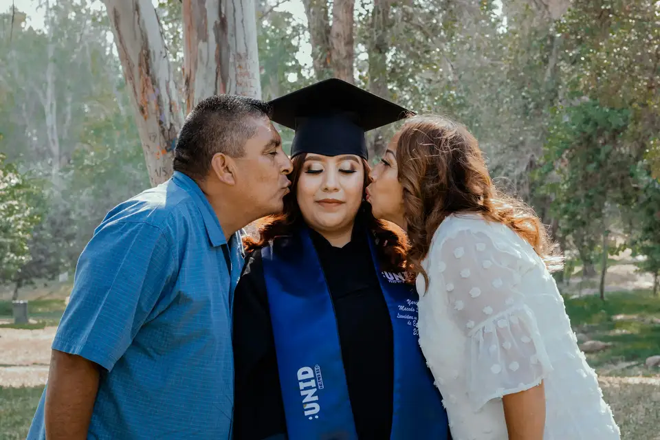 A graduate in her gown and blue stole gets a kiss on each cheek from her dad and her mom