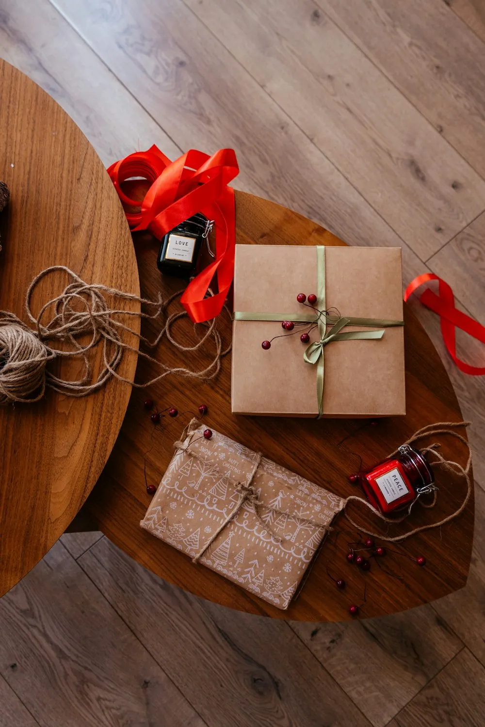 Regalos envueltos en papel kraft con lazo verde, un ovillo de cordel, cinta roja y velas sobre dos mesas de madera, vistos desde arriba
