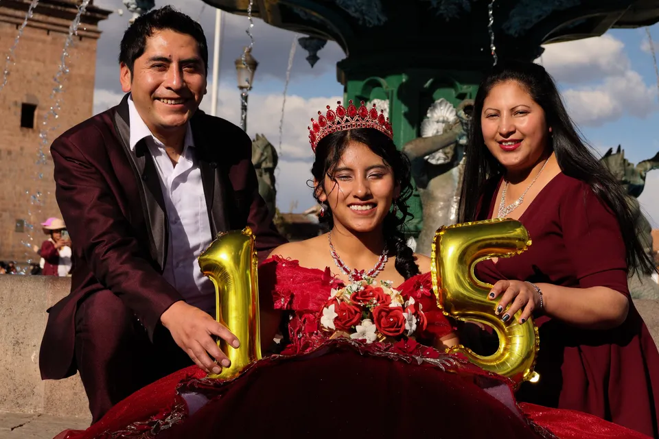The quinceañera, in a crown and a red gown, between her parents holding number 15 balloons, in Cusco